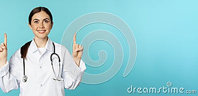 Enthusiastic medical worker, young woman doctor in white coat, stethoscope, showing advertisement, pointing fingers up Stock Photo