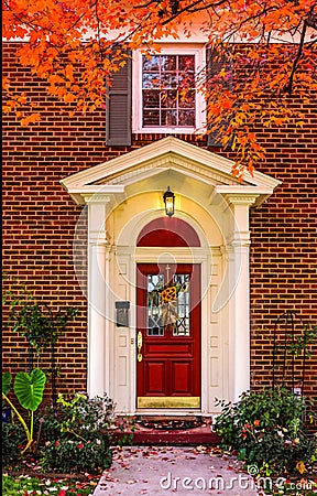 Entrance to brick house with pillars for porch and red door with autum leaves on sidewalk and colorful branches framing picture - Stock Photo