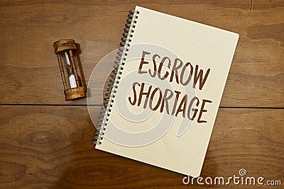 The entry Escrow shortage was made in a notebook with hourglass on wood table. Stock Photo