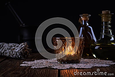Esoteric still life with burning candle, green vials with drops of alternative medicine and iron mortar on blurred black Stock Photo