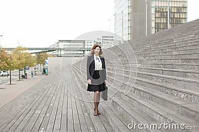 European businesswoman standing on stairs with bag and high buildings in background. Stock Photo
