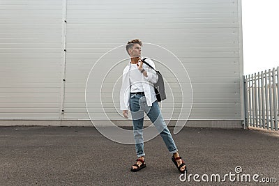 European young man model in stylish clothes in leather red sandals with a black bag is resting near the white modern building in Stock Photo