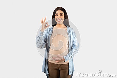 Everything is okay. Happy young pregnant lady gesturing okay and touching belly, looking and smiling at camera Stock Photo