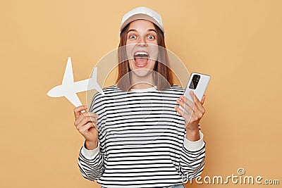 Excited amazed woman wearing striped shirt and baseball cap holding white paper plane and using smartphone booking tickets to Stock Photo