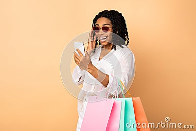 Excited Black Lady Using Phone Holding Shopper Bags, Beige Background Stock Photo