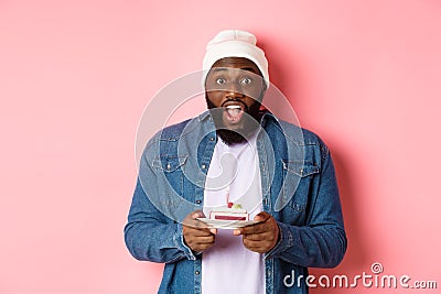 Excited Black man celebrating birthday, staring at camera amazed while making wish on bday cake with candle, standing Stock Photo