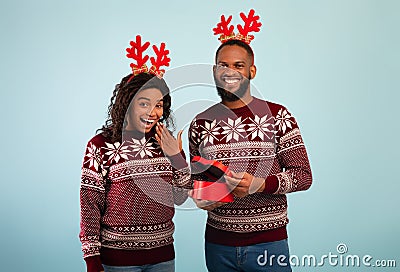 Excited black man giving wife Christmas gift, happy woman and her husband wearing deer horns, blue background Stock Photo