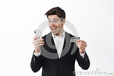 Excited businessman holding credit card, looking happy at smartphone screen, receive money transfer, standing over white Stock Photo