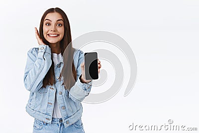 Excited, cheerful happy lively girl in denim jacket, jeans, holding smartphone, showing mobile phone screen as reacting Stock Photo