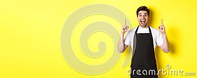 Excited coffee shop owner in black apron pointing fingers up, showing special offers, standing over yellow background Stock Photo