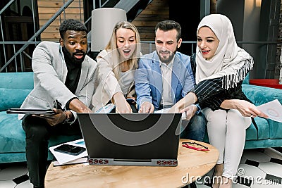 Excited group of diverse multiracial coworkers looking at the laptop screen with open mouth and smile, while sitting on Stock Photo