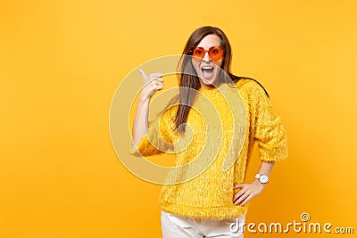 Excited happy young woman in fur sweater and heart orange glasses pointing thumb aside on copy space isolated on bright Stock Photo