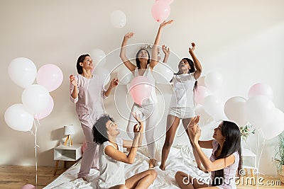 Excited multiracial girls throwing balloons in air at pajamas party. Stock Photo