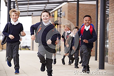 Excited primary school kids, wearing school uniforms and backpacks, running on a walkway outside their school building, front view Stock Photo