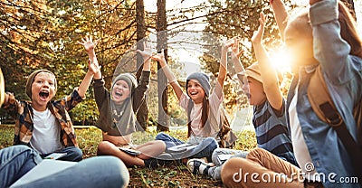 Excited school kids raising arms and screaming while sitting on grass in forest with notebooks Stock Photo