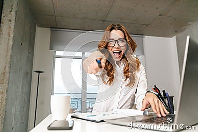 Excited young business woman dressed in formal clothes shirt indoors using laptop computer pointing to you Stock Photo
