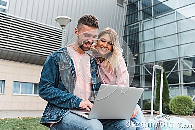 Excited young loving couple is waiting for flight at airport. Man is sitting and using laptop Stock Photo