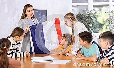 Excited young woman teacher showing national flag of France and telling preteens schoolchildren history of country Stock Photo