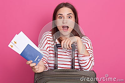 Excited young woman in white shirt with red stripes keeping mouth open, expresses astonish, holds pass port and boarding pass Stock Photo