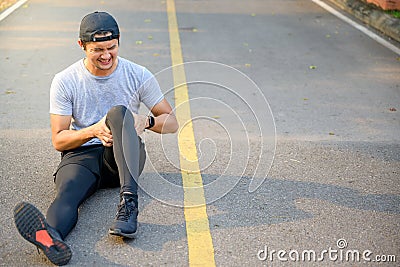 Exercise Injury Concept: A young Asian runner sits down on a park street due to pain in his knee and uses a hand to grasp the sore Stock Photo