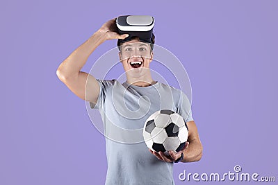 Exercising with virtual reality concept. Young athletic guy in VR headset holding soccer ball on lilac studio background Stock Photo