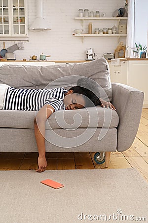 Exhausted African American woman sleeping on couch at home, dropping cellphone on the floor. Fatigue Stock Photo