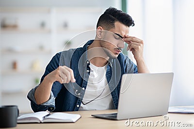 Exhausted arab guy sitting at workdesk, removing glasses Stock Photo