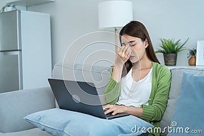 Exhausted Asian business woman hurt eye while using laptop computer. Young girl office worker sit on sofa feel visual fatigue and Stock Photo