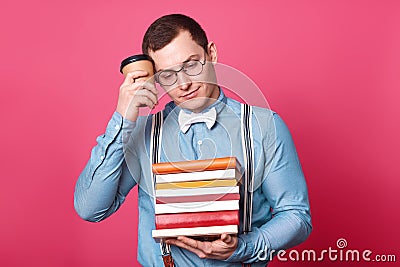 Exhausted athletic student touches his temple with papercup of drink, looks aside, holds books in one hand, having lunch during Stock Photo