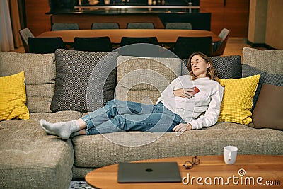 Exhausted fatigued young woman napping lying on sofa in living room, sleeping with phone after hard day at home Stock Photo