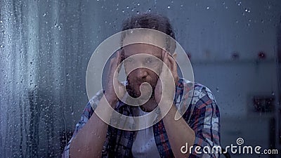Exhausted middle-aged man massaging temples behind rainy window, migraine pain Stock Photo