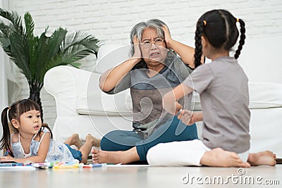 Exhausted old elderly grandmother sit on floor in living room and feel unwell tired from little children running and playing loud Stock Photo