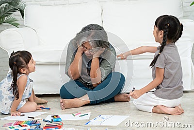 Exhausted old elderly grandmother sit on floor in living room and feel unwell tired from little children running and playing loud Stock Photo