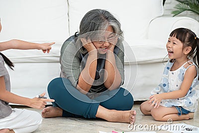 Exhausted old elderly grandmother sit on floor in living room and feel unwell tired from little children running and playing loud Stock Photo