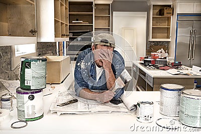 Exhausted and tired painter with face in hands holding wet paint brush in messy home kitchen with paint cans everywhere Stock Photo