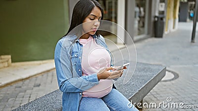 Expecting young woman, casually sitting on city bench, emotionally touching her pregnant belly while texting on smartphone, Stock Photo