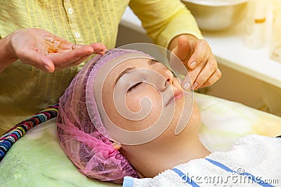 An experienced cosmetologist applies a mask of emulsion on the face of a young girl who lies on the couch during facial cleansing Stock Photo