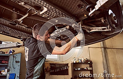 Experienced young car mechanic standing under lifted car during repair process in garage. Car check up and maintenance Stock Photo