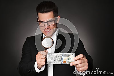 Expert authenticating 100 dollar banknote with magnifying glass against dark background, focus on hand. Fake money Stock Photo