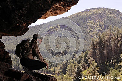 EXPLORER WITH HAT SUNGLASSES AND BACKPACK RESTING AT THE ENTRANCE OF A CAVE WATCHING THE MOUNTAINS Stock Photo