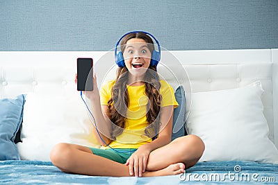 Expressive emotional excited teen girl showing screen phone. Teenager child girl wearing headphones listening music on Stock Photo