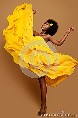Expressive Woman dancing in Yellow Flying Dress. Happy Dark Skinned Dancer in Waving Fabric Gown. Model with Black curly Afro Hair Stock Photo