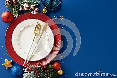 Top view featuring elegant plates, golden flatware, wine glass, ornaments, candle, confetti, frosty fir branches on blue backdrop Stock Photo