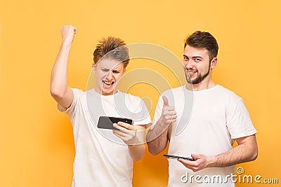 Extreme men emotionally play games on a smartphone, stand on a yellow background, wear white t-shirts. Two people are emotionally Stock Photo