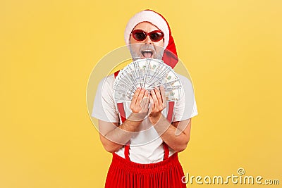 Extremely excited happy gray bearded man in santa claus hat and stylish sunglasses holding fan of dollars, giving money on Stock Photo