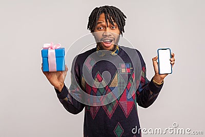 Extremely surprised pleased african guy with dreadlocks holding mobile phone with empty blank and gift box, online presents order Stock Photo