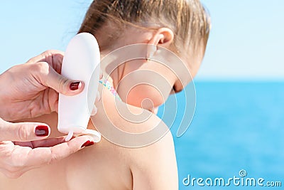 Eyropean mom is taking sun protector cream from white tube for applying it at shoulder of her young pretty daughter on the beach c Stock Photo