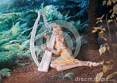 Fabulous forest elf sits under tree and plays on white harp, girl with long blond hair braided in long yellow dress Stock Photo