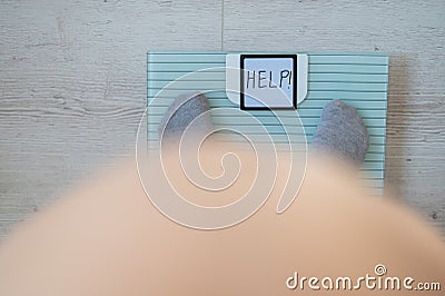 Faceless fat woman measures weight on a bathroom scale. Top view of a large naked female belly and feet in gray socks on Stock Photo