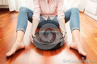 Faceless middle section of young woman seated at floor holding an intelligent vacuum cleaner to clean the floor Stock Photo